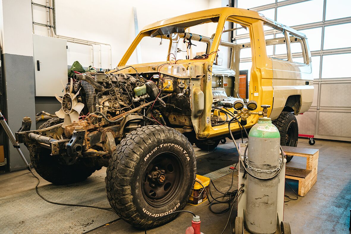 Ein gelber Geländewagen im Rahmen der Restauration bei AllRoad. Der Motorraum ist freigelegt, und Werkzeuge sowie Gasflaschen stehen für die Reparatur bereit. 