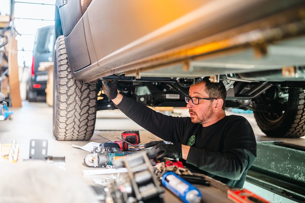 Ein Mechaniker von AllRoad überprüft den Unterboden eines Campers in der Werkstatt. Umgeben von Werkzeugen und Ersatzteilen inspiziert er präzise das Fahrwerk und den Unterflur des Fahrzeuges. 