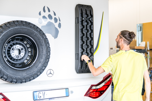 Ein zufriedener Kunde von AllRoad begutachtet die neuen Sandbleche an seinem HYMER ML-T570 in unserer Service Werkstatt für Wohnmobile. Ein Reserverad mit Ersatzradträger von DesertService und das Mercedes-Logo sind ebenfalls sichtbar.