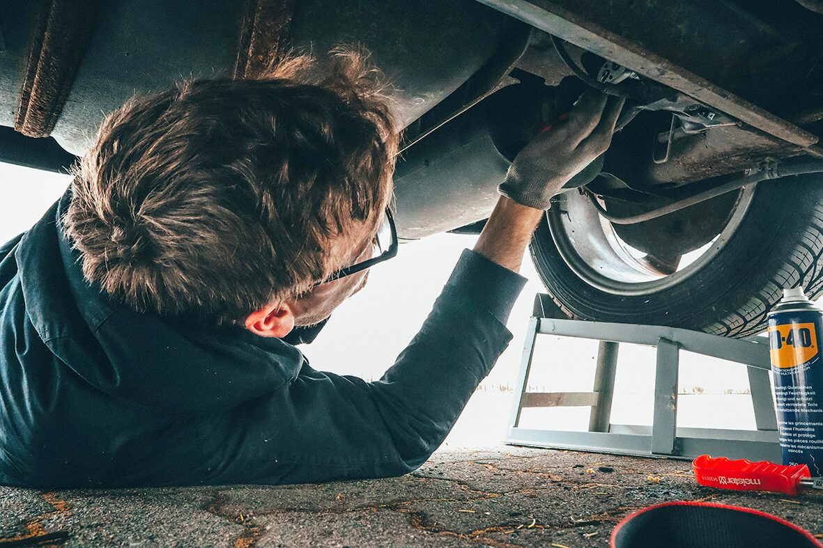 Ein Mechaniker von AllRoad arbeitet am Unterboden eines Fahrzeugs. Werkzeuge und Schmiermittel sind griffbereit neben ihm positioniert. Hier findet die Fehlersuche direkt am Fahrzeug statt. 